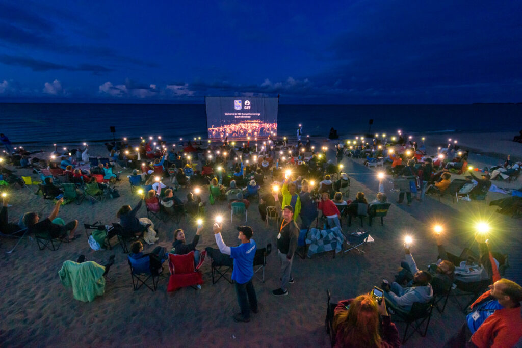 A crowd sits on a beach in front of a movie screen set against Lake Winnipeg on a dark evening, holding their phone flashlights into the air.