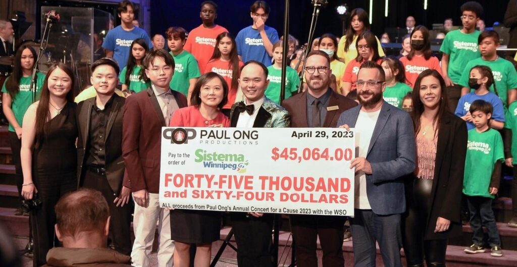 Five people stand at the front of a stage holding a large cheque reading "Forty-five thousand and sixty-four dollars" made out to Sistema Winnipeg from Paul Ong's Annual Concert for a Cause 2023 with the WSO.