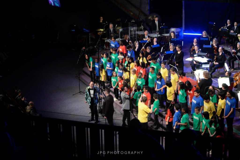 A large group of people wearing green, blue, red and yellow shirts stand on a stage.