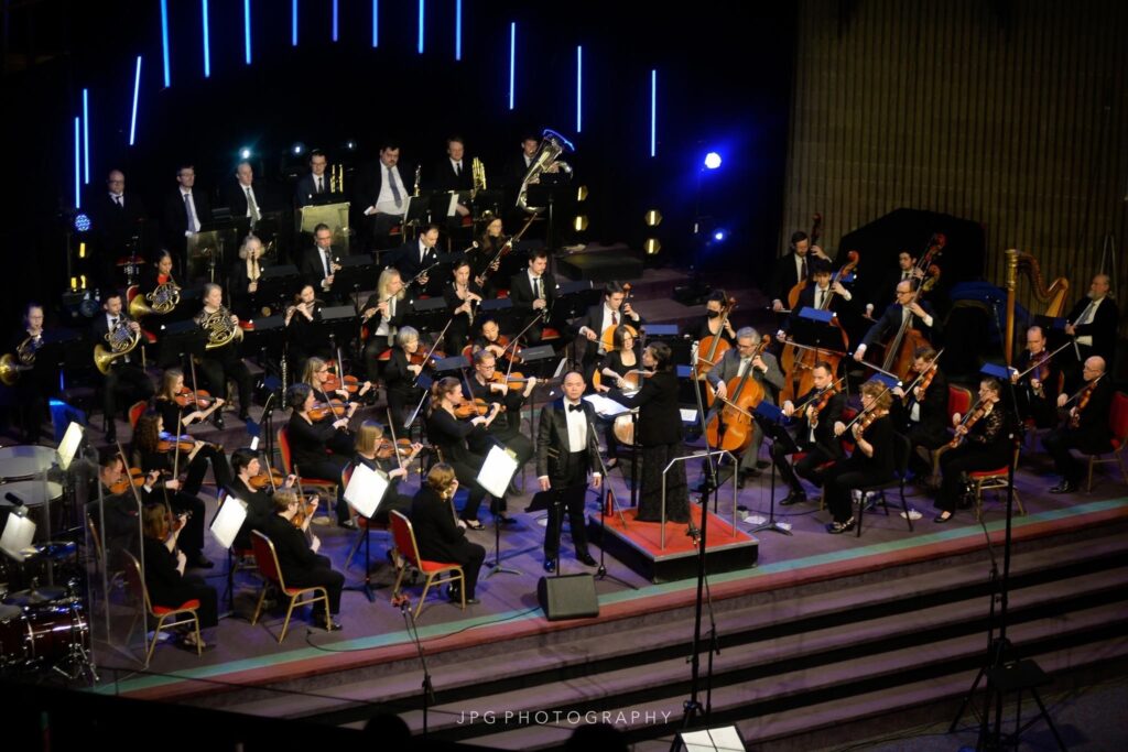 A man in a tuxedo stands at the front of a stage surrounded by a symphony orchestra.