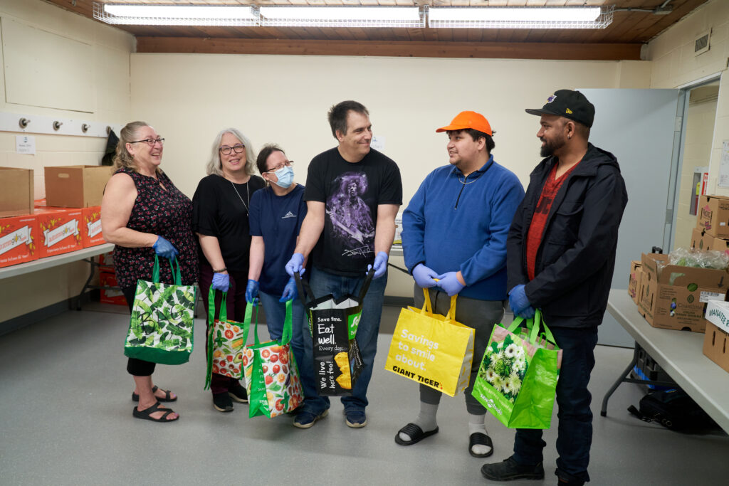 A group of volunteers stand in a row smiling, holding colourful bags to be filled with vegetables, in a small room lined with tables.