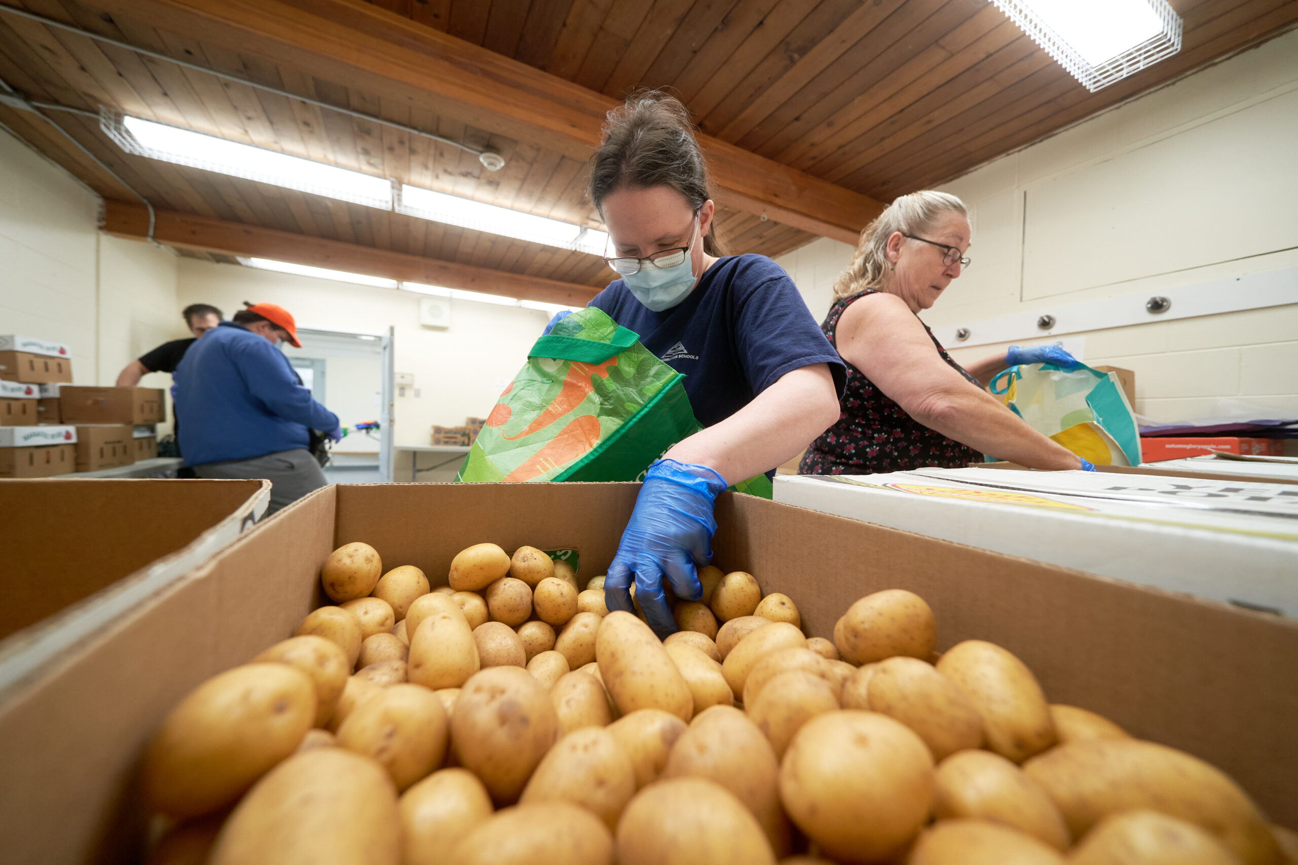 A woman in a blue shirt wearing glasses and a mask reaches a gloved hand into a box of potatoes.