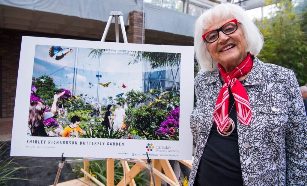 A woman with white hair, red glasses and a red scarf stands beside a rendering of the Shirley Richardson Butterfly Garden.