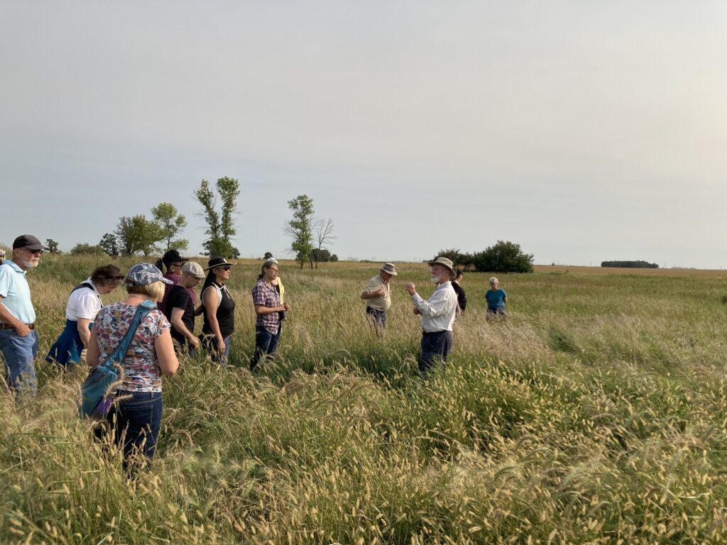 A man with a white beard and khanki-coloured wide-brimmed hat stands among tall-grass prairie surrounded by a small audience.