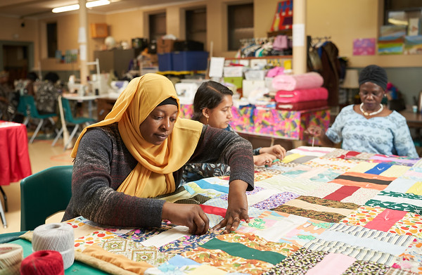 Three women sit at a table piecing together colourful pieces of fabric into a quilt.