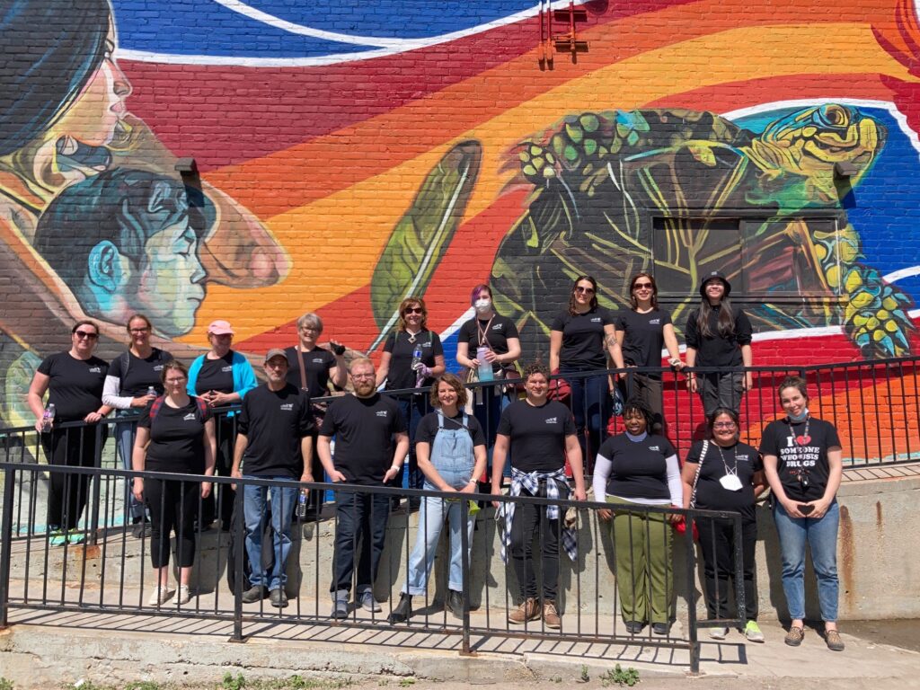 A group of people wearing black t-shirts stand on a ramp in front of a colourful mural and wave at the camera.