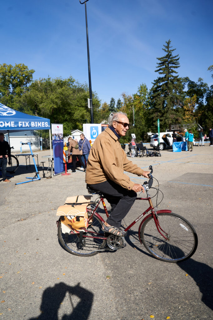 A man with sunglasses and a tanned jacket rides a bike in front of a mobility fair for older adults.