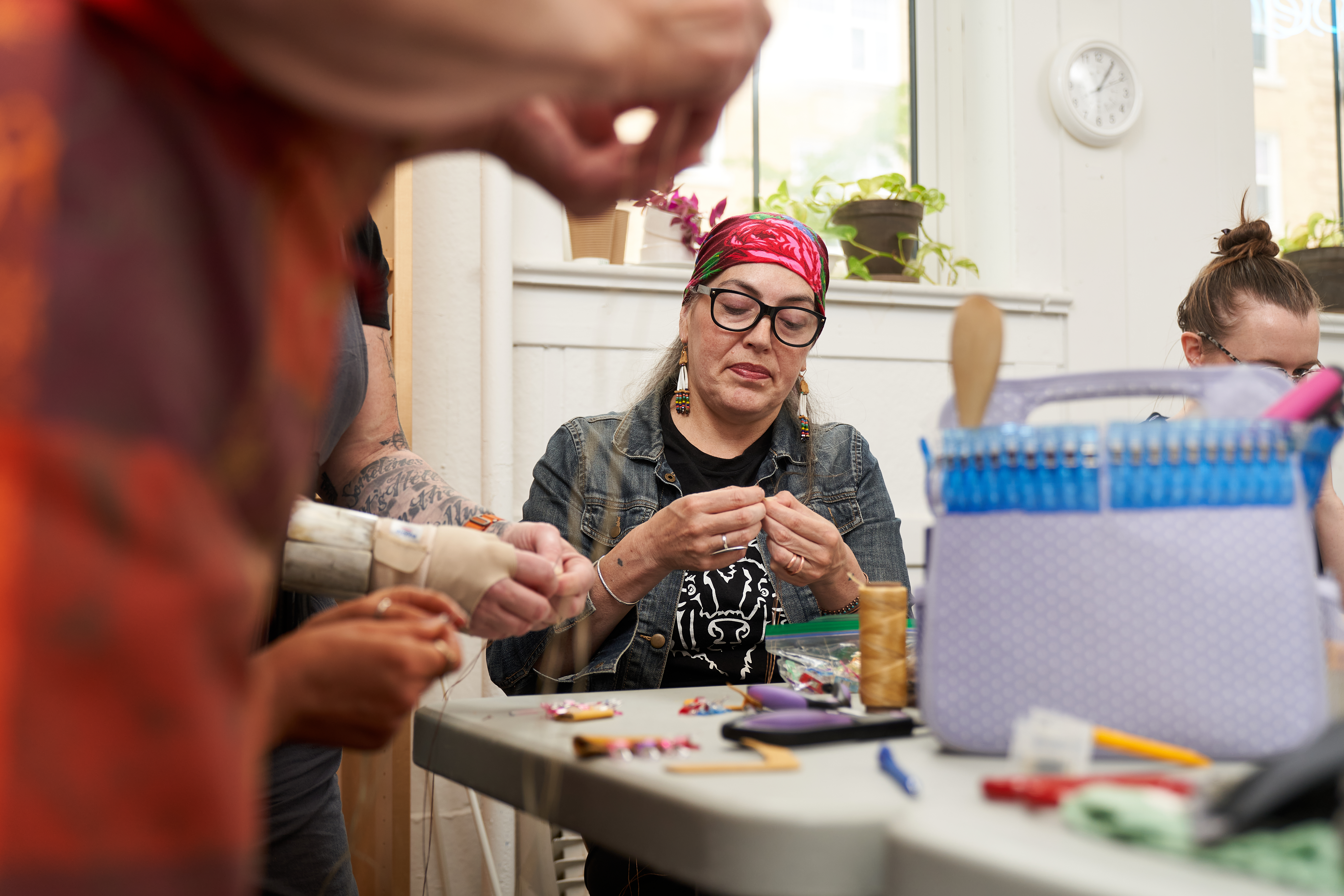 A woman with glasses, a bandana and long earrings works on a pair of mittens.