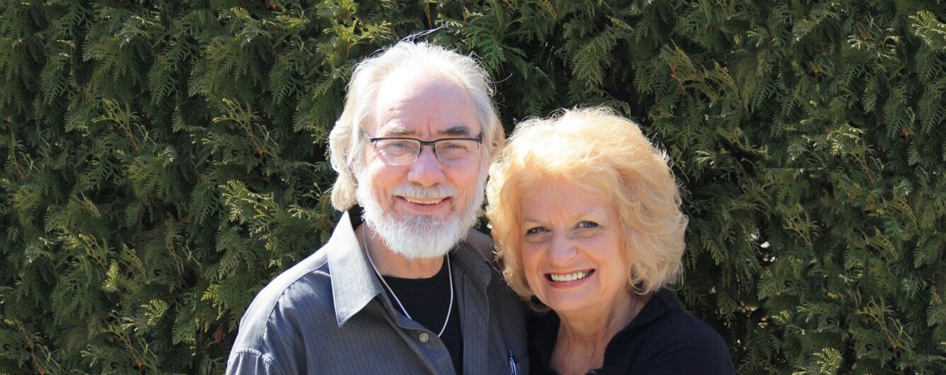 Wilma and Cliff Derksen smile together outdoors in front of tall green trees.