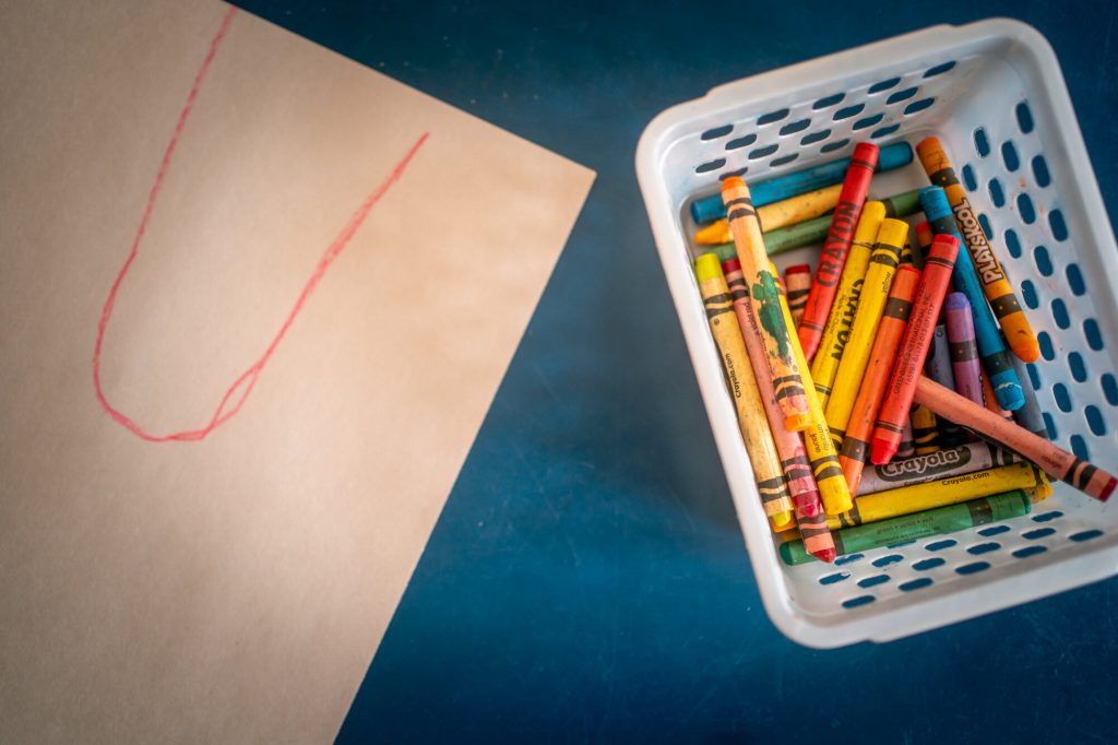 A basket of crayons sits beside a child’s drawing on a blue classroom table.