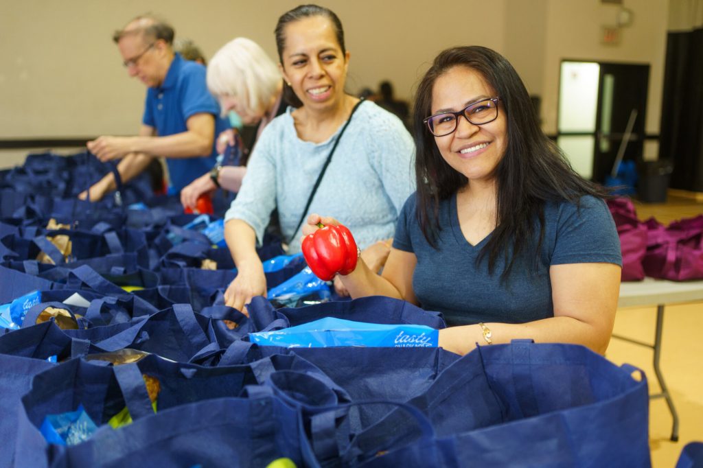 A smiling woman holds a red pepper while helping organize grocery bags at a community food distribution event.