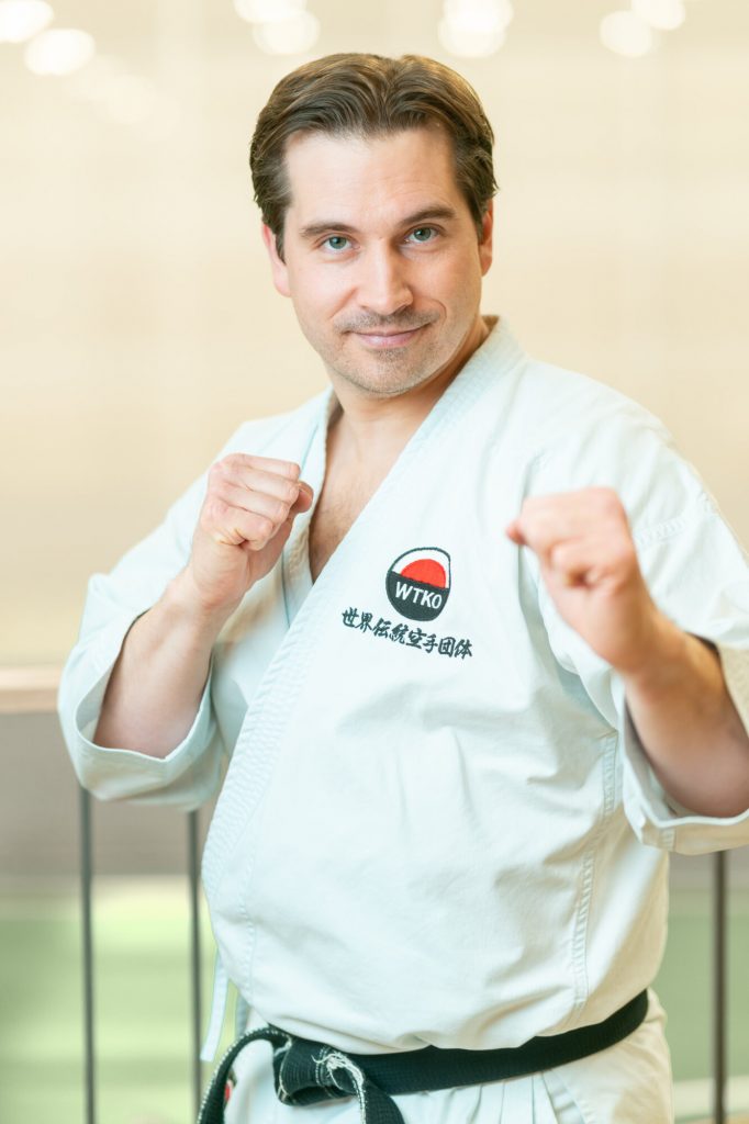 A karate instructor in a white gi and black belt poses with raised fists in a fighting stance.
