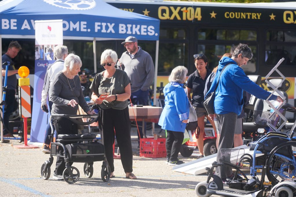 People gather outdoors at a community accessibility fair while browsing mobility equipment and community resource booths.