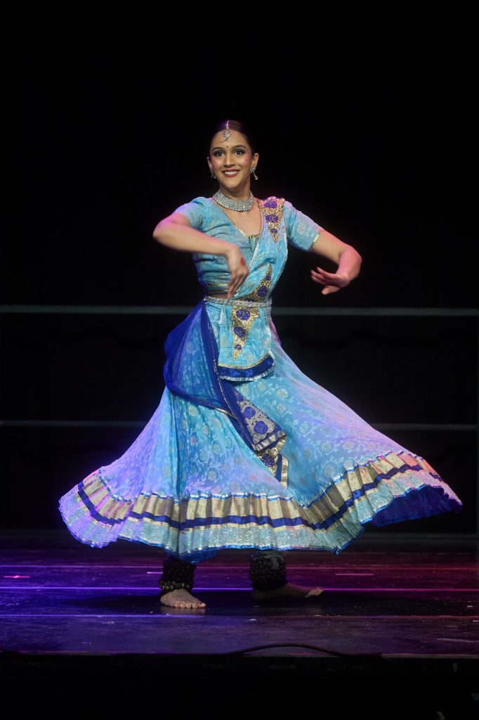 A dancer in a blue traditional outfit performs on stage under theatre lighting.