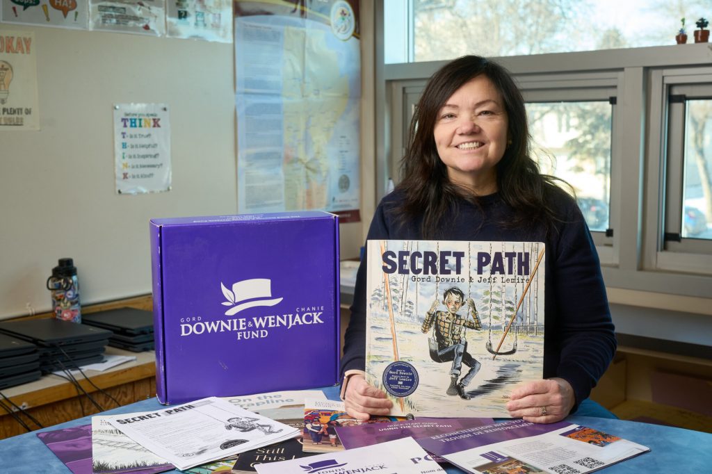 A woman smiles while holding a copy of Secret Path beside educational resources from the Gord Downie & Chanie Wenjack Fund.