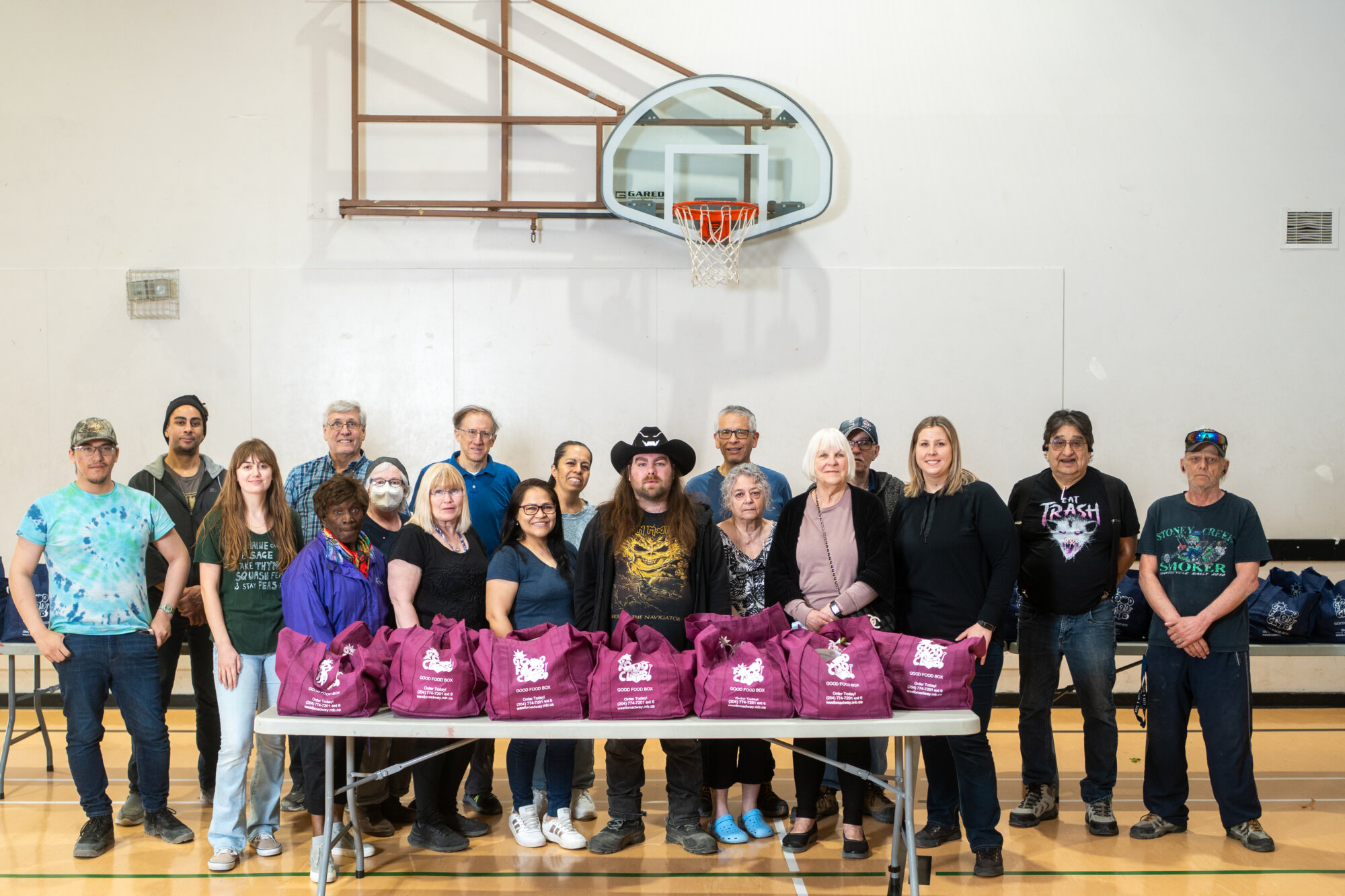 Group of community members standing in a gymnasium behind a table filled with purple Good Food Club bags.
