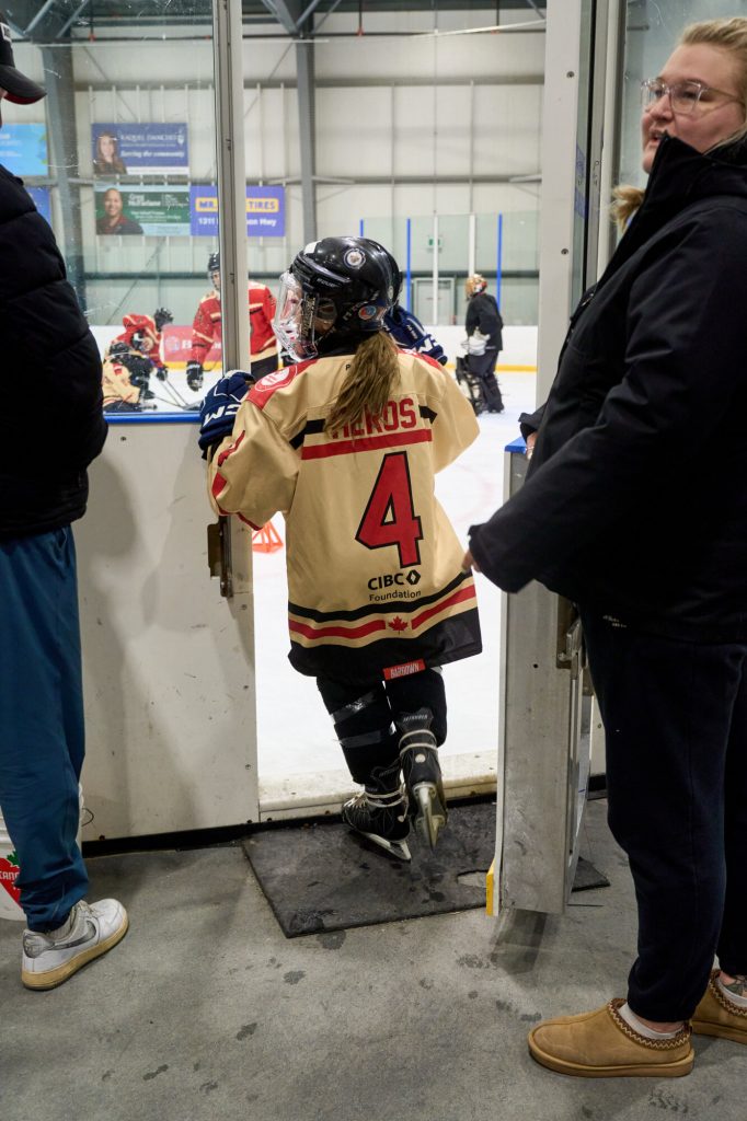 A young hockey player wearing a HEROS jersey enters the ice rink while coaches and participants stand nearby.
