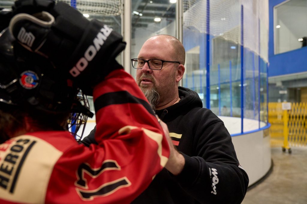 A hockey coach adjusts a young player’s equipment beside the rink boards during practice.