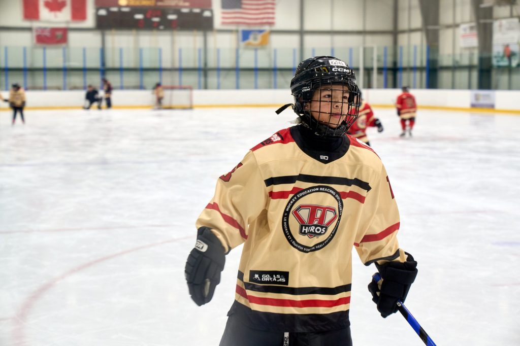 A young hockey player in a cream-coloured HEROS jersey stands on the ice holding a hockey stick.