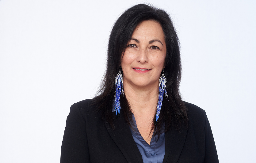 Professional headshot of a smiling woman wearing a black blazer and blue dress shirt against a neutral light grey background.