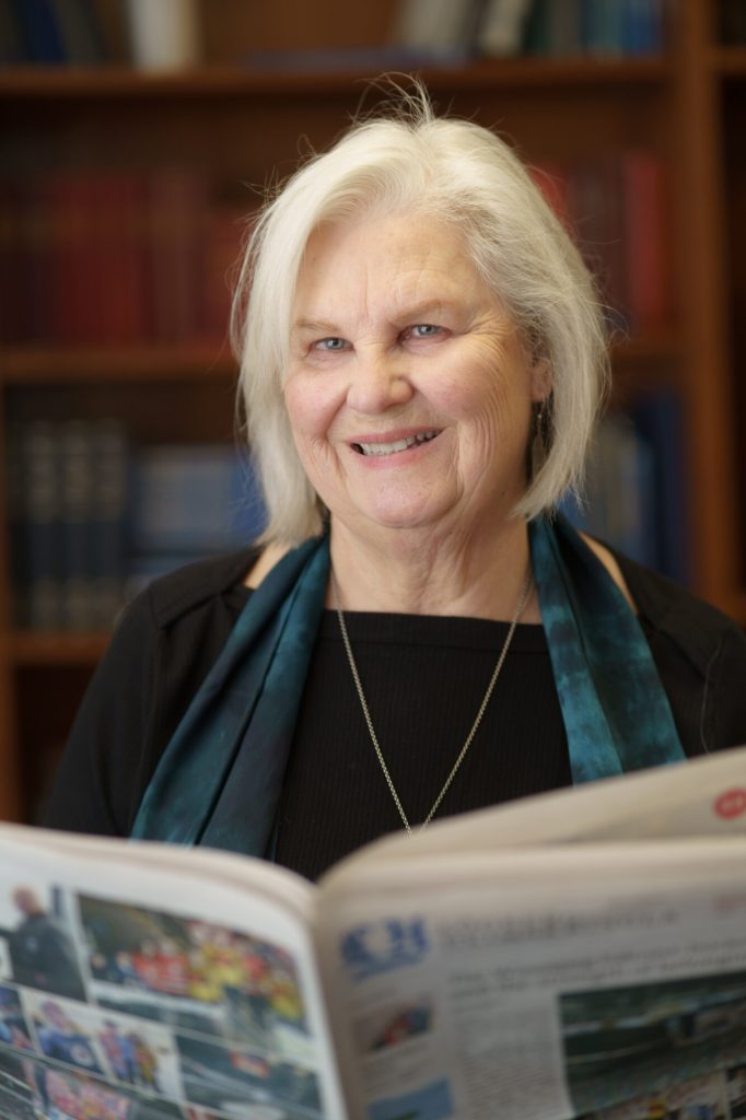 A woman smiles while holding a copy of the Lögberg-Heimskringla newspaper.