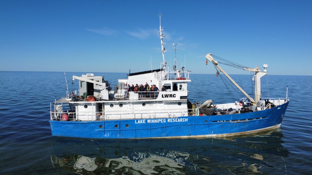 A blue-and-white Lake Winnipeg Research vessel marked “LWRC” floats on calm open water under a clear blue sky, with several people gathered on deck and the boat reflected in the lake.