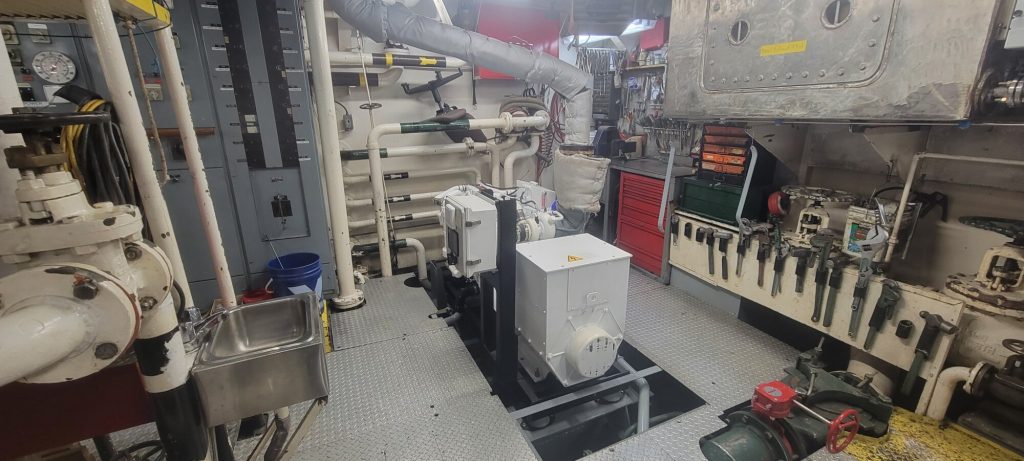 A new generator installed inside the MV Namao’s engine room, surrounded by pipes, control panels, tools, and mechanical equipment.