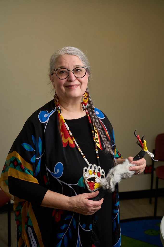 A woman wearing beaded earrings and a patterned shawl holds a ceremonial item.
