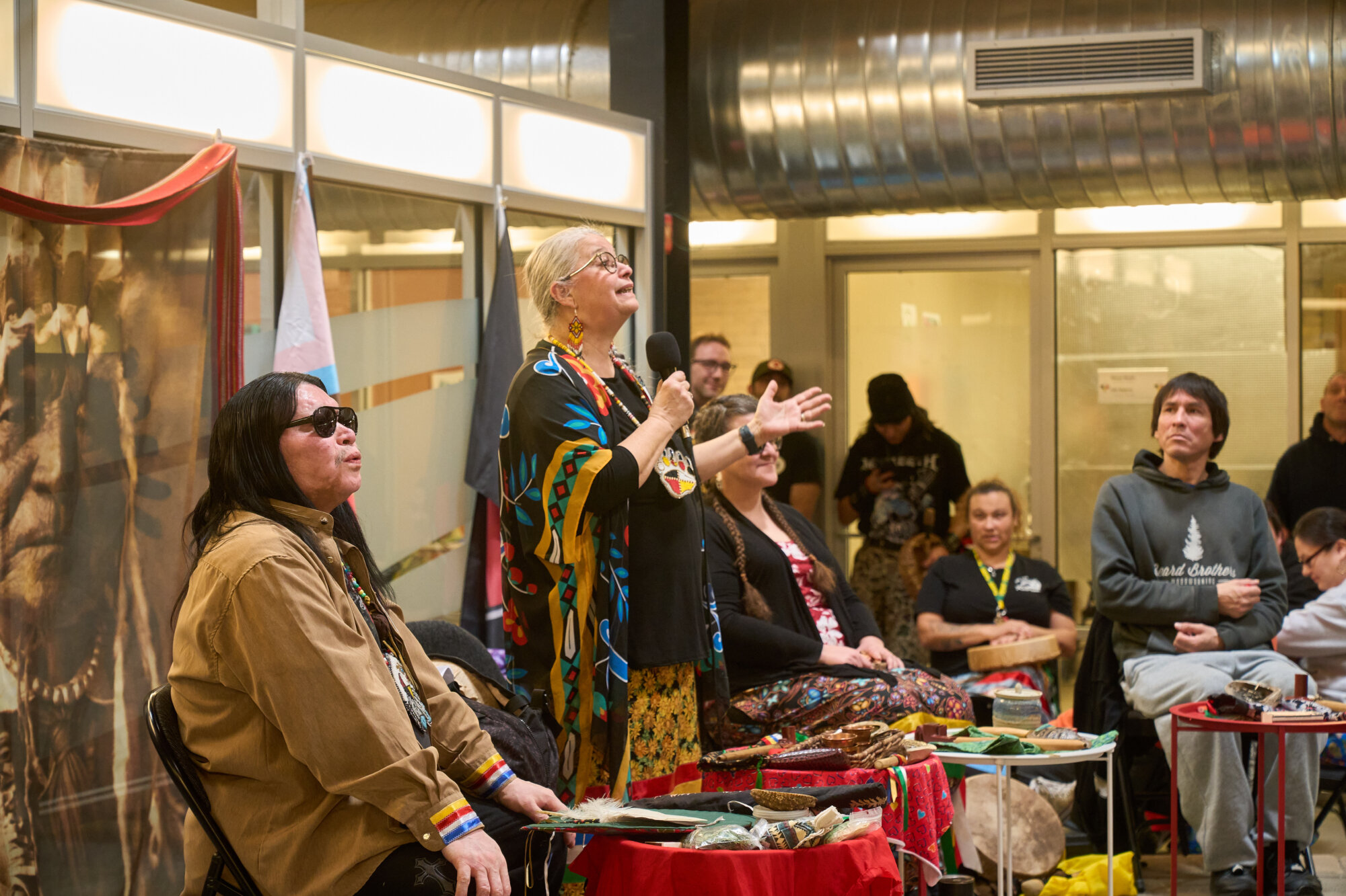 An Indigenous speaker addresses a group gathered indoors while seated participants and audience members listen during a cultural gathering.