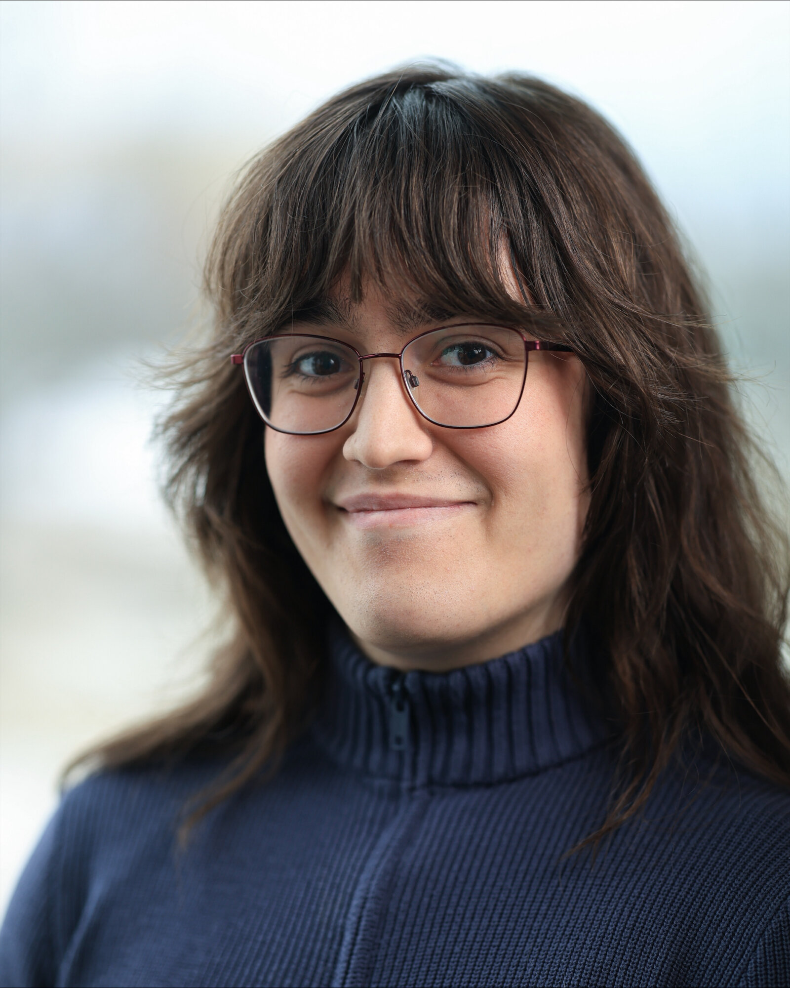 Portrait of volunteer award recipient Mae Junke smiling at the camera, wearing glasses and a blue ribbed sweater against a soft, light background.
