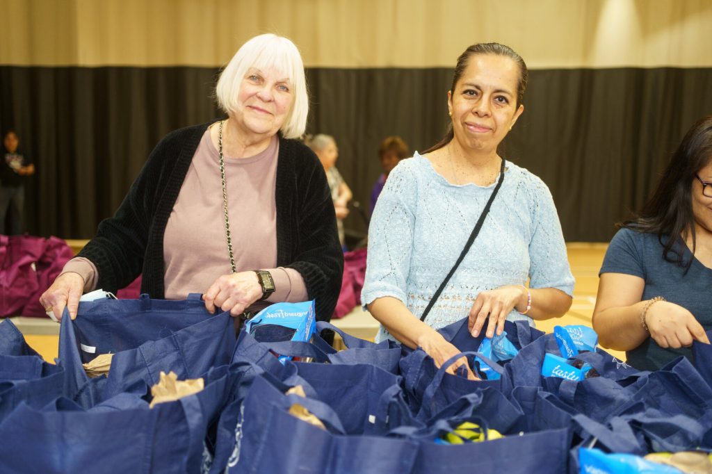 Two women stand behind tables filled with blue grocery bags during a community food distribution event.