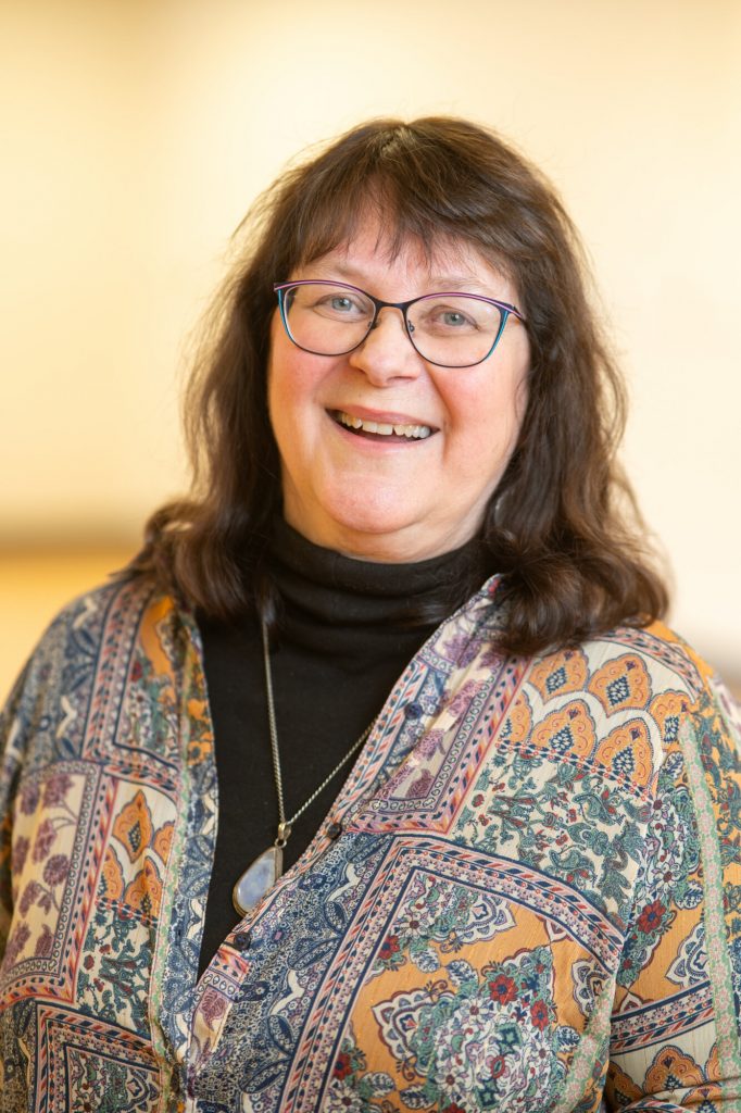 A smiling woman wearing glasses and a patterned blouse stands indoors.