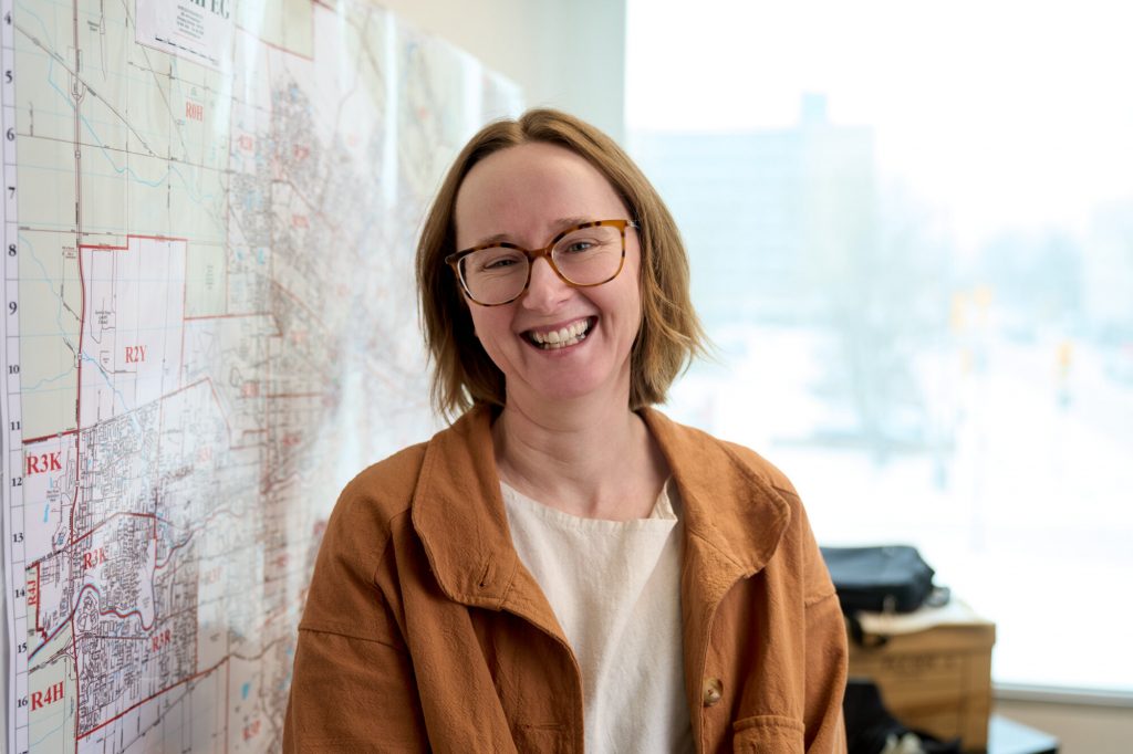 A smiling woman wearing glasses and a brown jacket stands beside a large map inside the New Journey Housing office.
