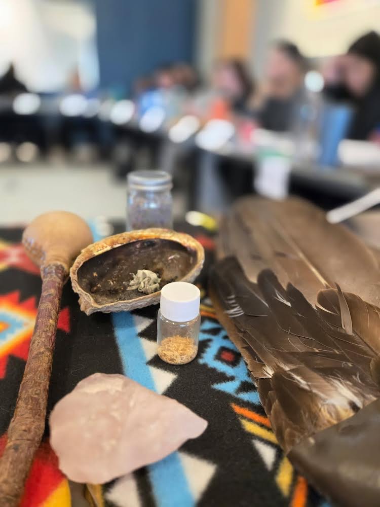 Ceremonial items including feathers, medicines, and a shell are displayed on a patterned blanket during a workshop.