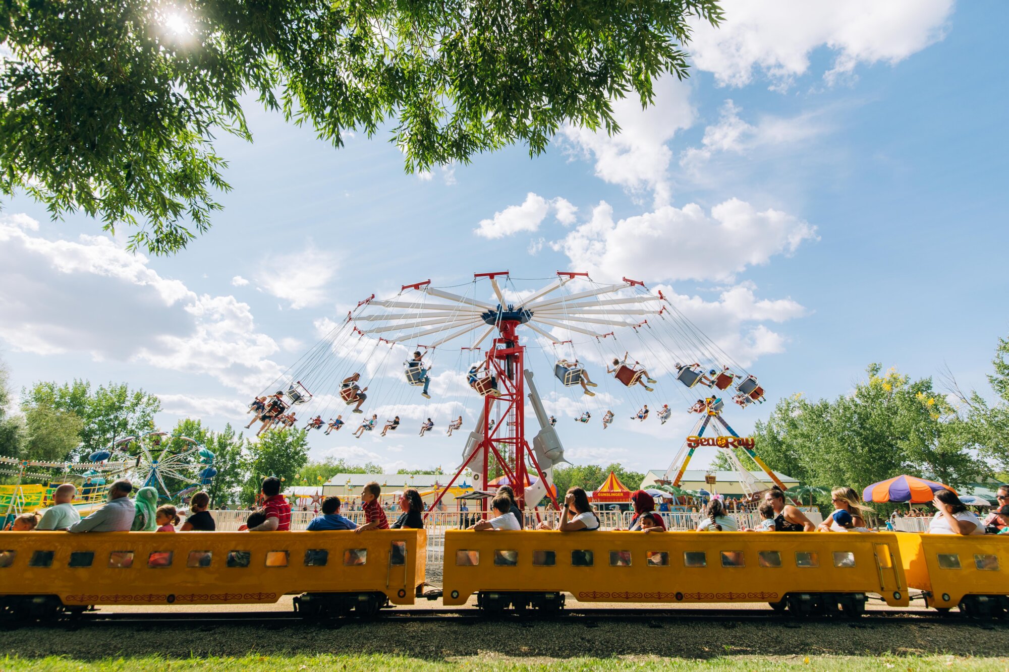 Families ride a miniature train in front of a large swing ride at an outdoor fair on a sunny summer day.