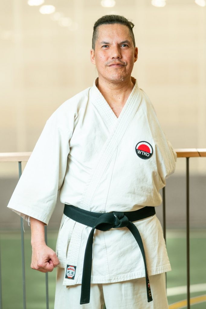 A karate instructor wearing a white gi and black belt stands in a training space.