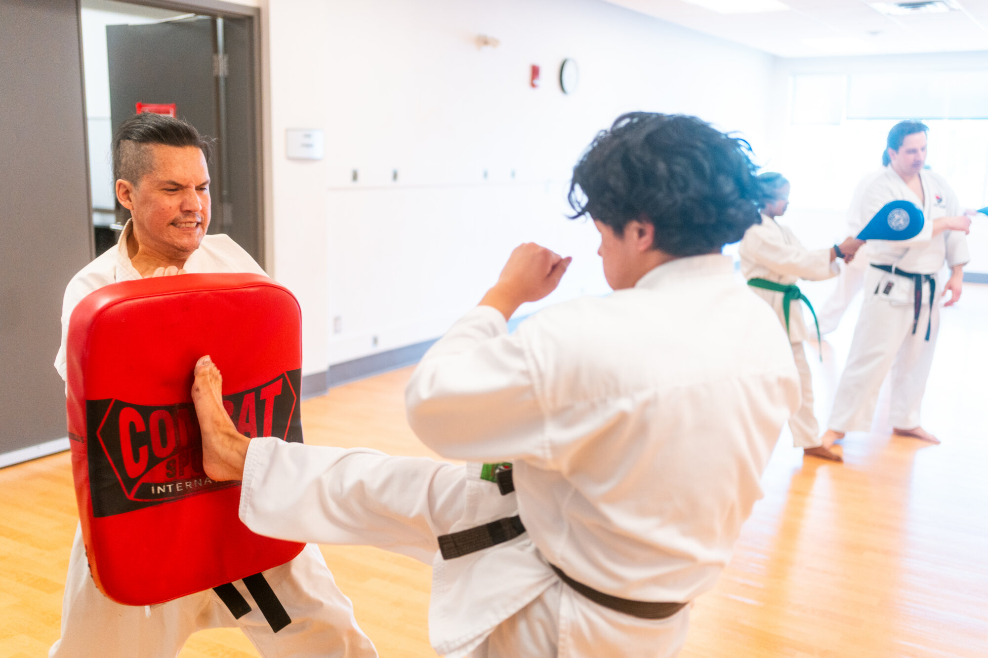 A student practices a kick on a red training pad held by an instructor.