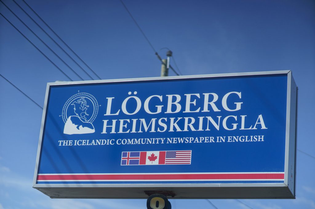 A blue roadside sign for Lögberg-Heimskringla, an Icelandic community newspaper in English.