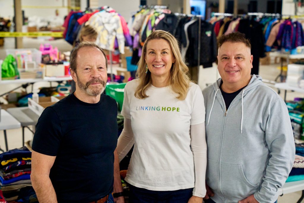 Rob Crowley, Kristie Pearson and Lawrence “Spatch” Mulhall stand smiling in the Linking Hope warehouse, surrounded by tables and racks of sorted donated clothing and supplies.
