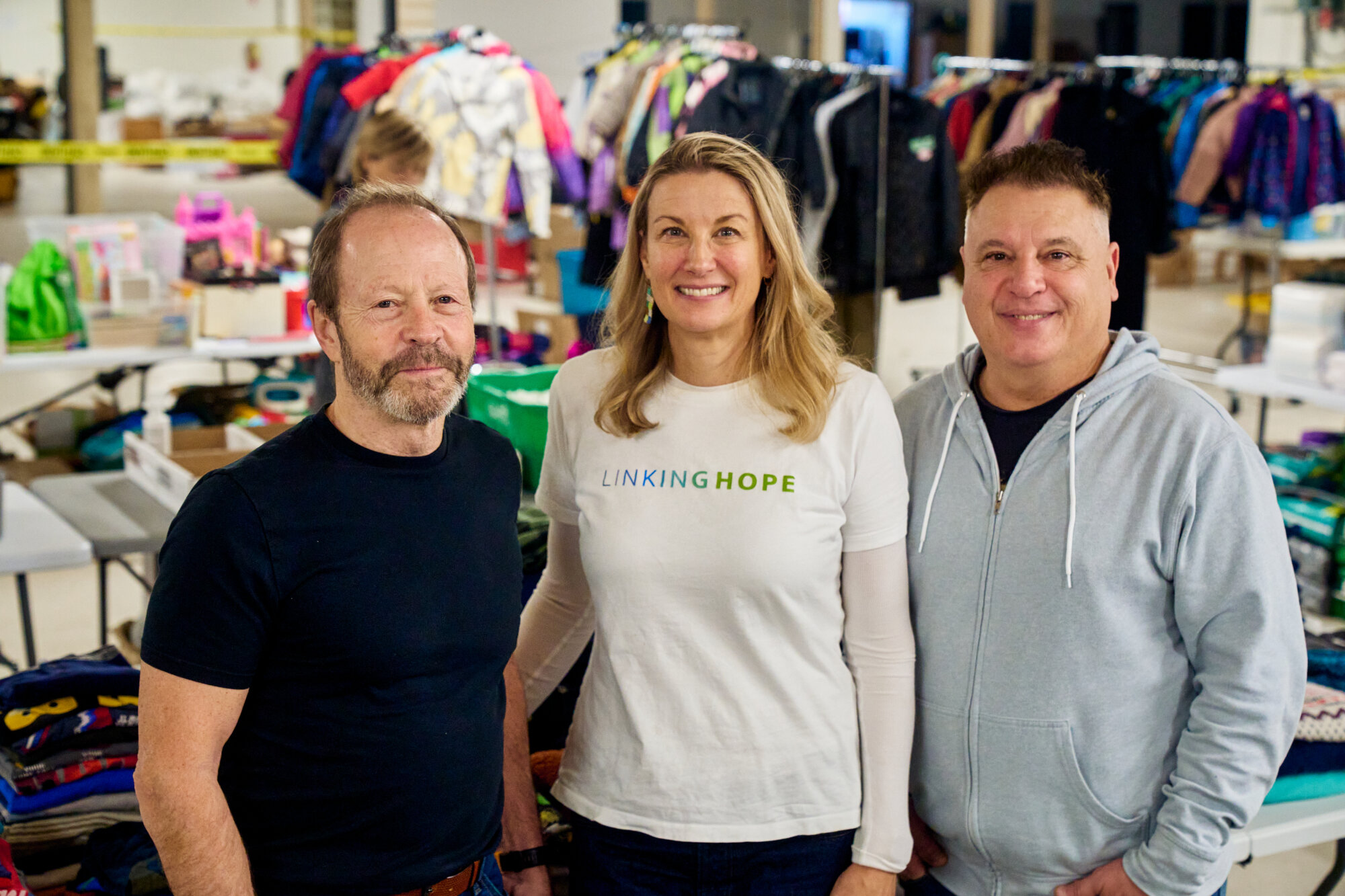 Rob Crowley, Kristie Pearson and Lawrence “Spatch” Mulhall stand smiling in the Linking Hope warehouse, surrounded by tables and racks of sorted donated clothing and supplies.