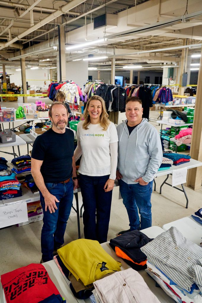 Rob Crowley, Kristie Pearson and Lawrence “Spatch” Mulhall stand smiling in the Linking Hope warehouse, surrounded by tables and racks of sorted donated clothing and supplies.
