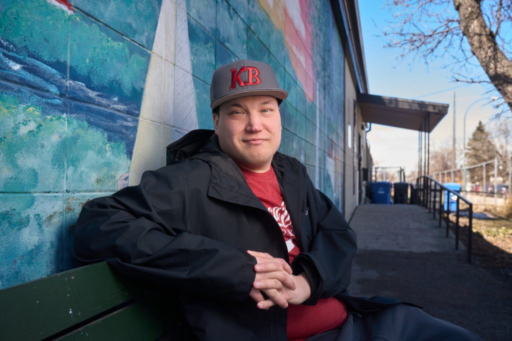 A man wearing a black jacket and grey “KB” baseball cap sits on a bench beside a colourful mural outside a community building.