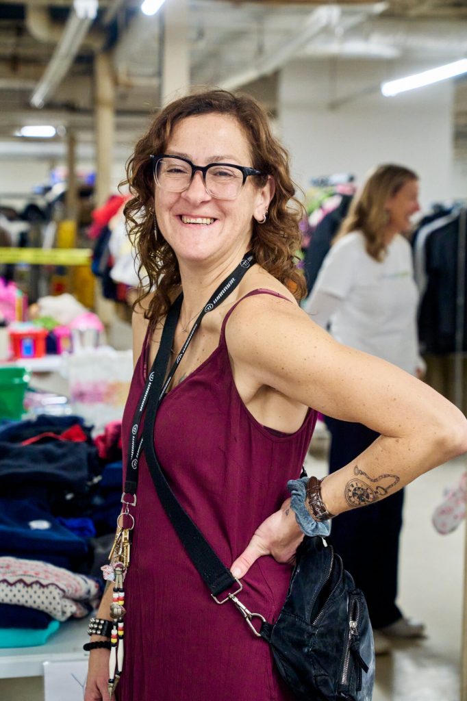 Tara Bell, Agency & Volunteer Support Worker at Linking Hope, stands in the organization’s warehouse surrounded by sorted clothing and essential items.