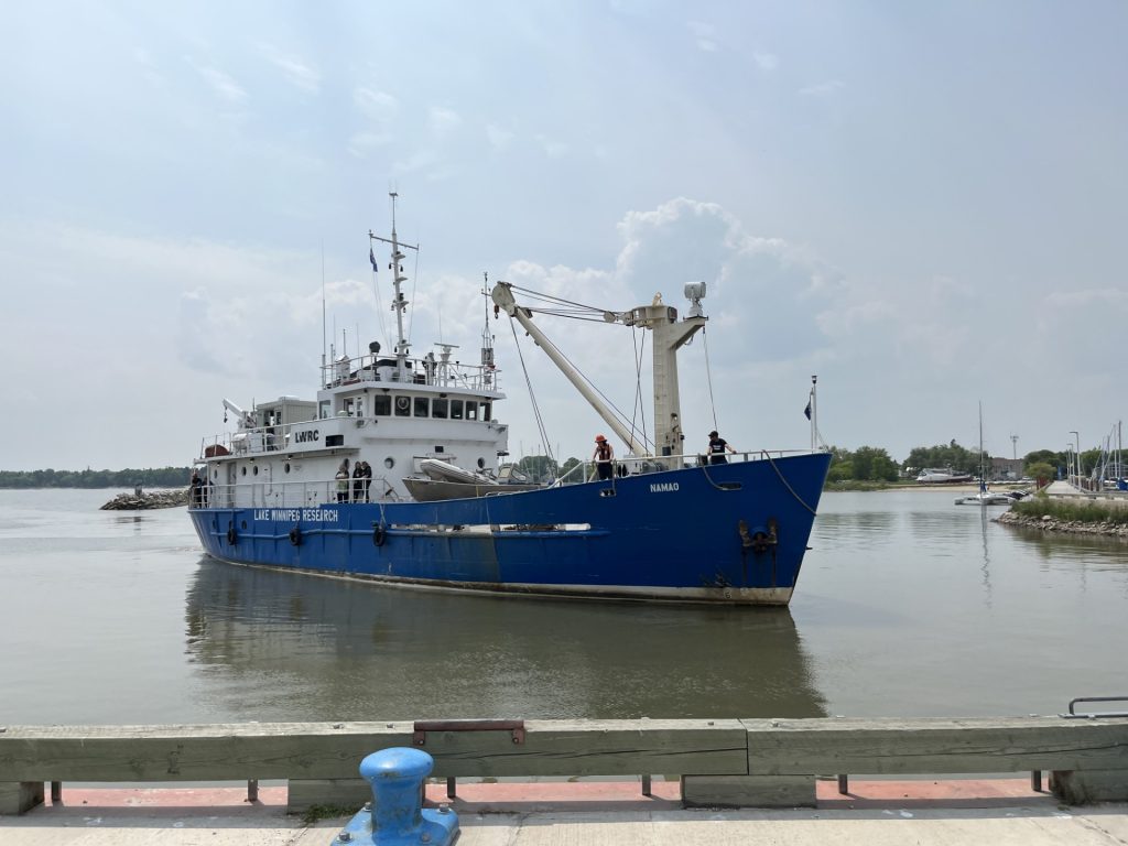 The blue-and-white MV Namao research vessel docked in Gimli Harbour on Lake Winnipeg, with crew members on deck and calm water around the boat.